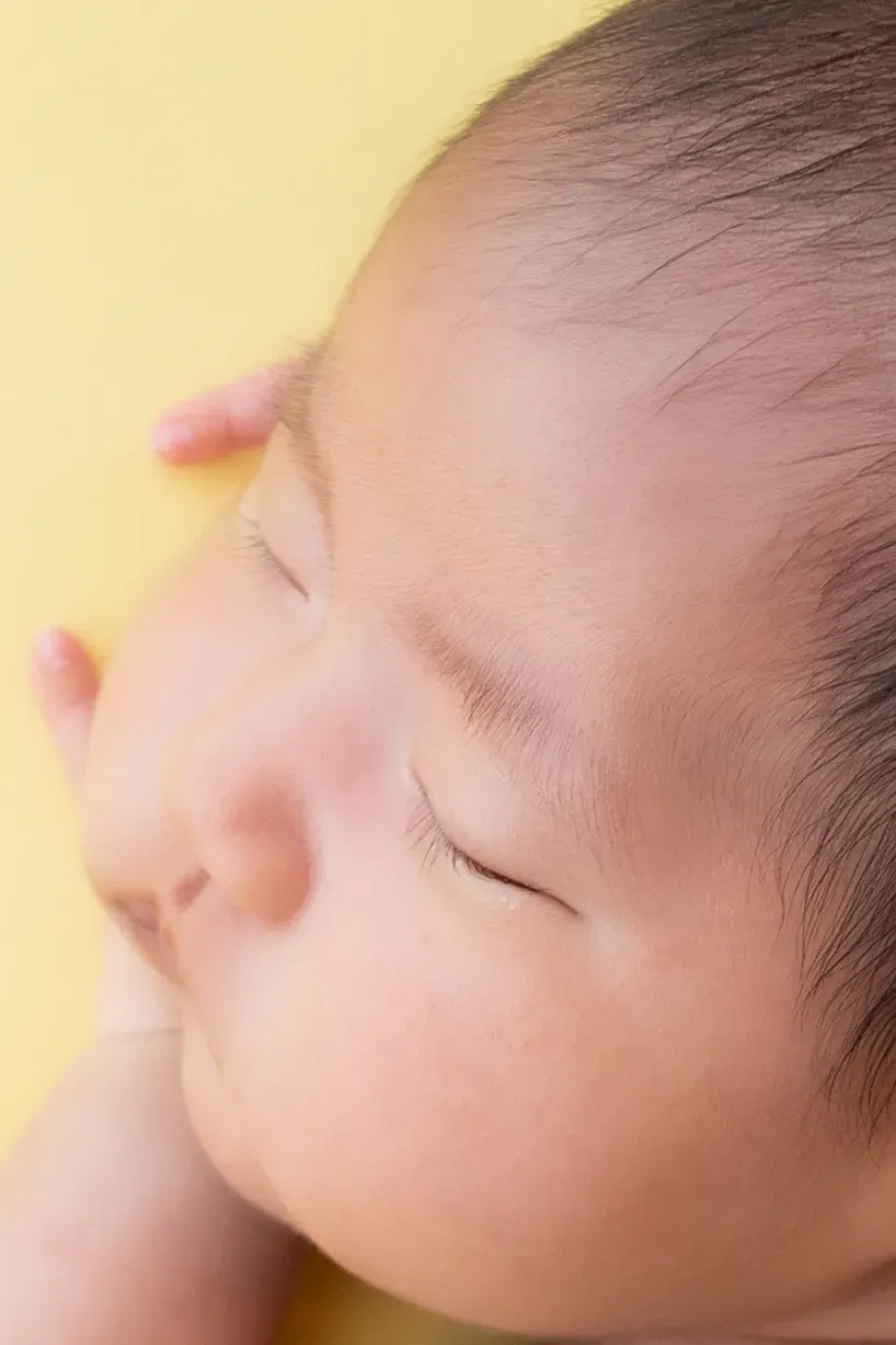 黄色い背景の上で、手を顔に添えて眠る赤ちゃんの横顔のアップ