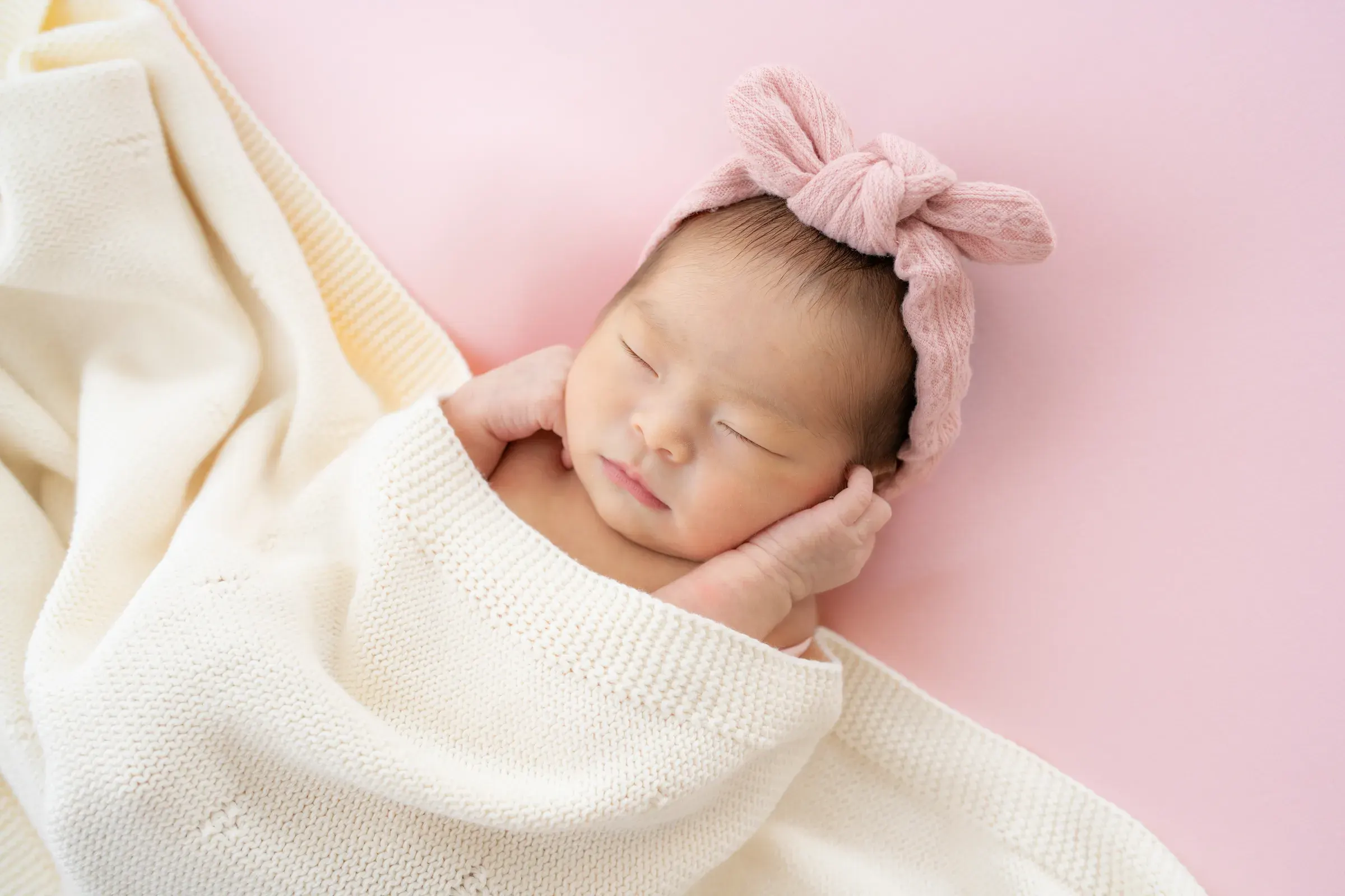 ピンクのリボンをつけ、両手を頬に添えて眠る赤ちゃん