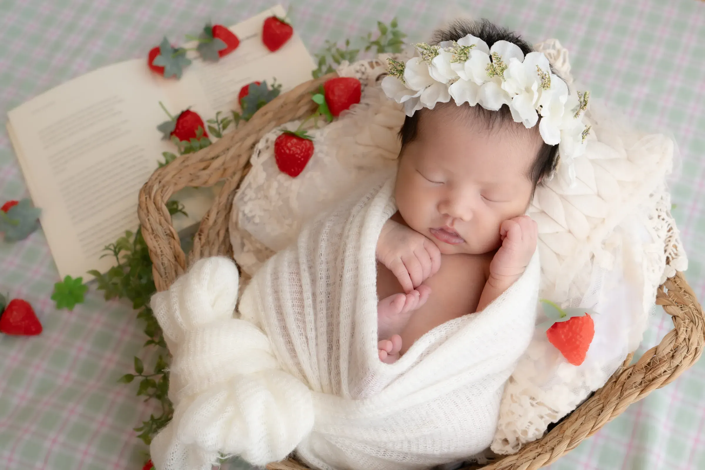 いちごと本を添えたチェック柄の上で、白いおくるみに包まれ籠の中で眠る赤ちゃん
