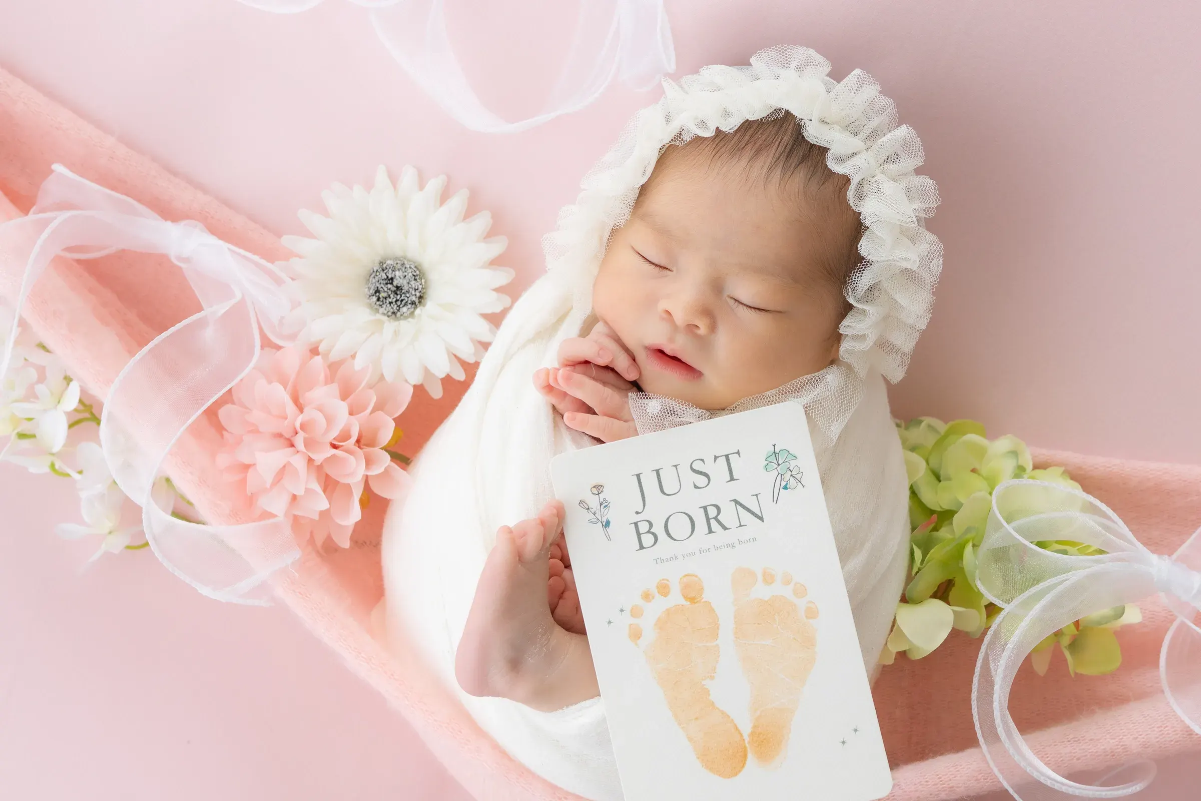ピンクの布とお花に囲まれ、「JUST BORN」の足形カードを抱いて眠る赤ちゃん
