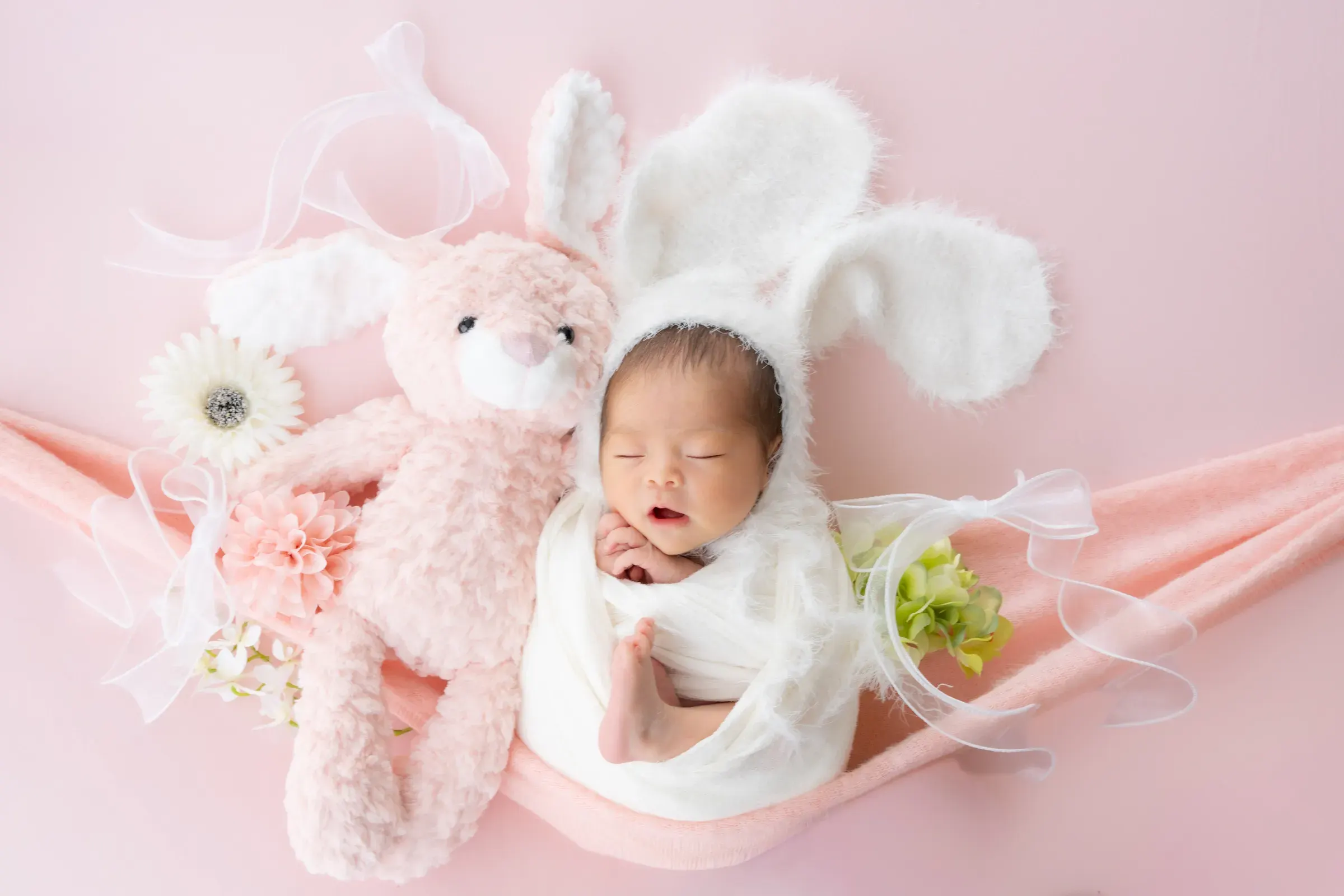 うさぎのぬいぐるみと並び、うさ耳の帽子で眠る赤ちゃん。ピンクの背景