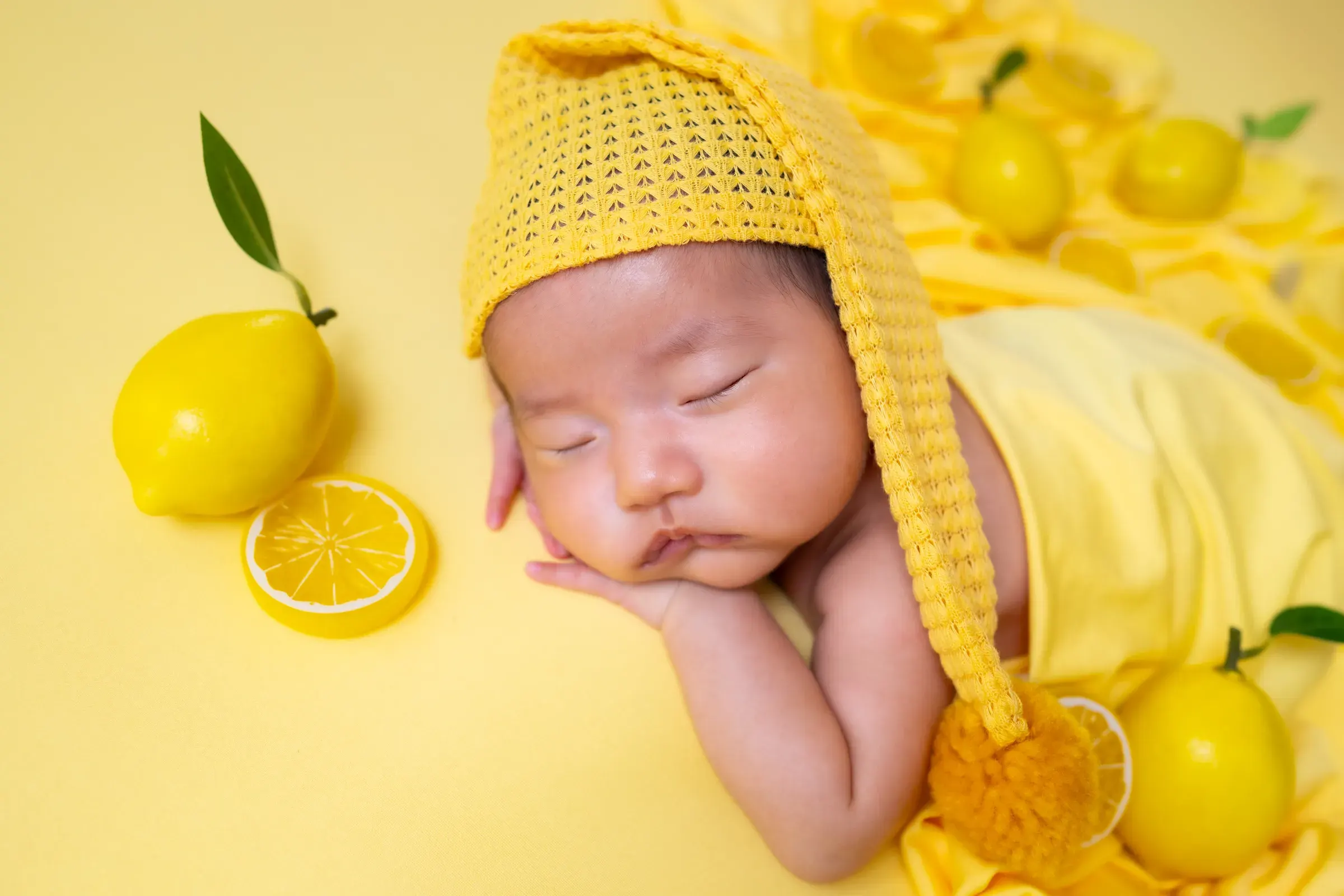 レモン色のニット帽をかぶり、頬杖をつくように眠る新生児。黄色い背景にレモンが並ぶ